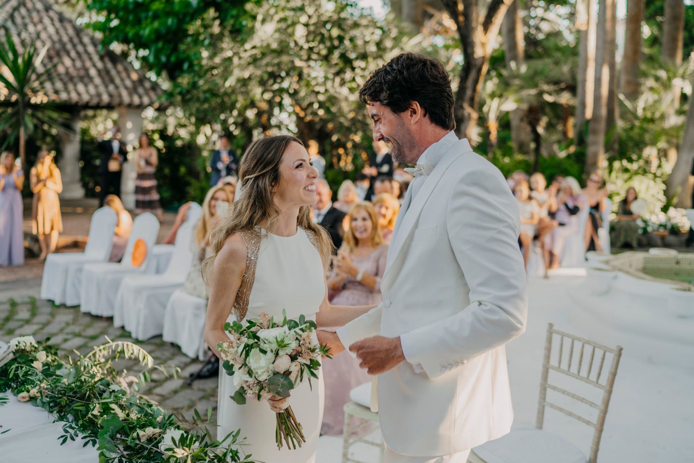 Couple shares a joyful dance moment outdoors during an elegant garden wedding ceremony with white decor and greenery.