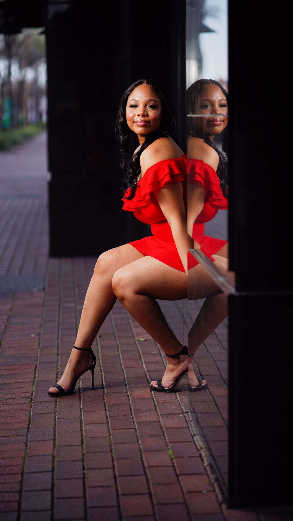 A person in a red off-shoulder dress poses elegantly against a reflective surface on brick pavement.