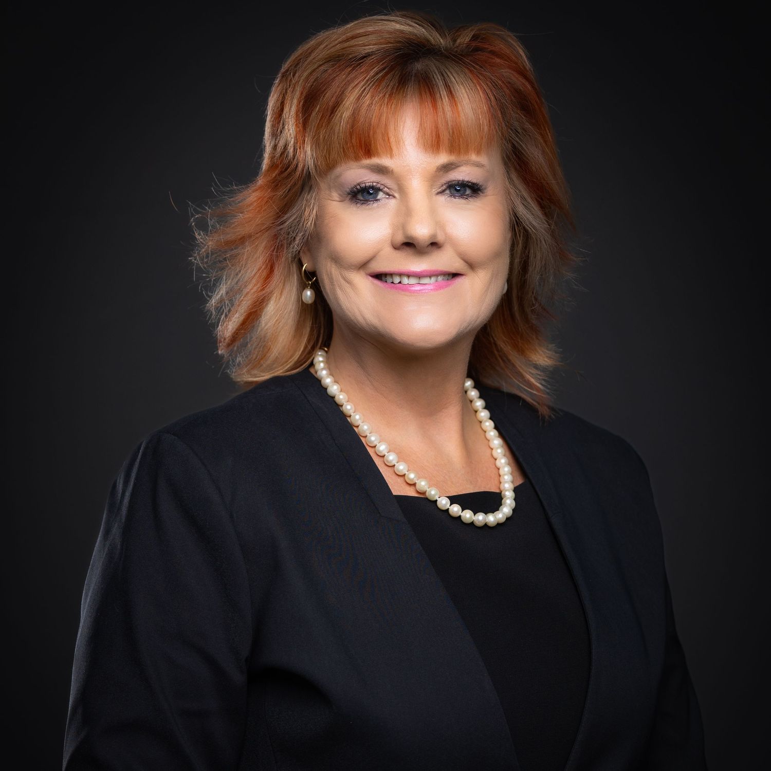 Professional headshot of a smiling woman wearing a black blazer and pearl necklace against a dark background.