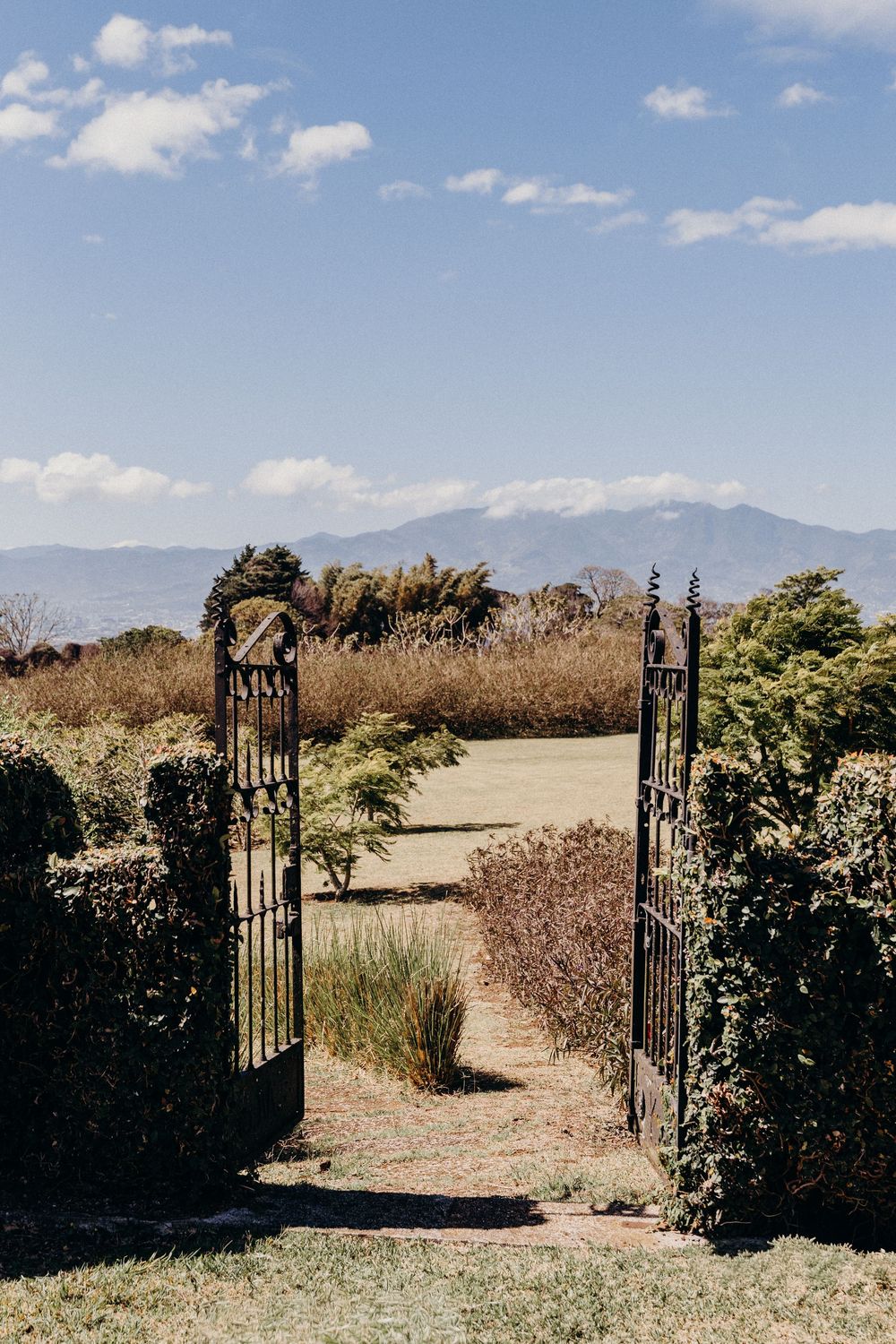Open wrought iron garden gate leading to a scenic landscape with mountains and blue sky in the background.