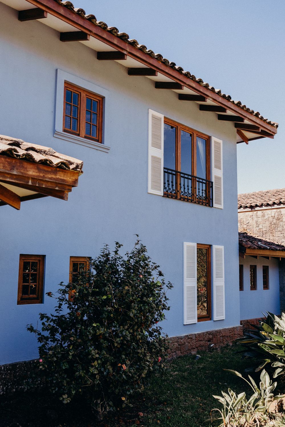 Blue two-story colonial house with wooden windows, white shutters, terracotta roof tiles, and lush green shrubs in foreground.