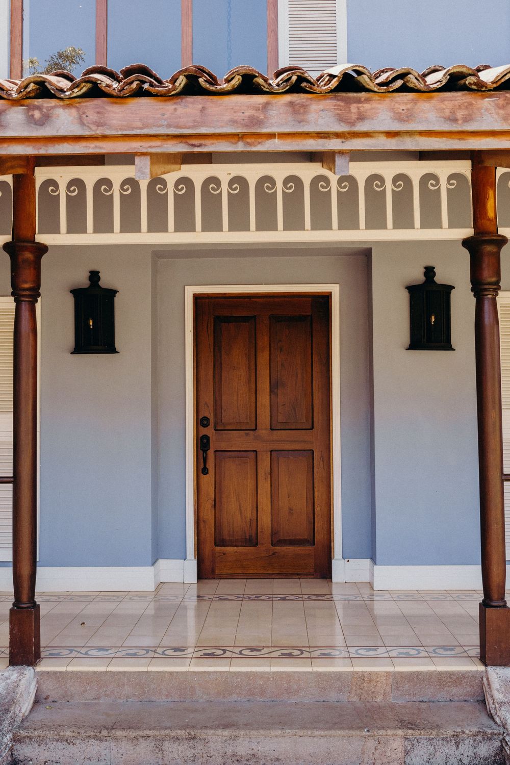 Elegant wooden front door with black lantern sconces on a blue stucco home with decorative cornice and tile roof.