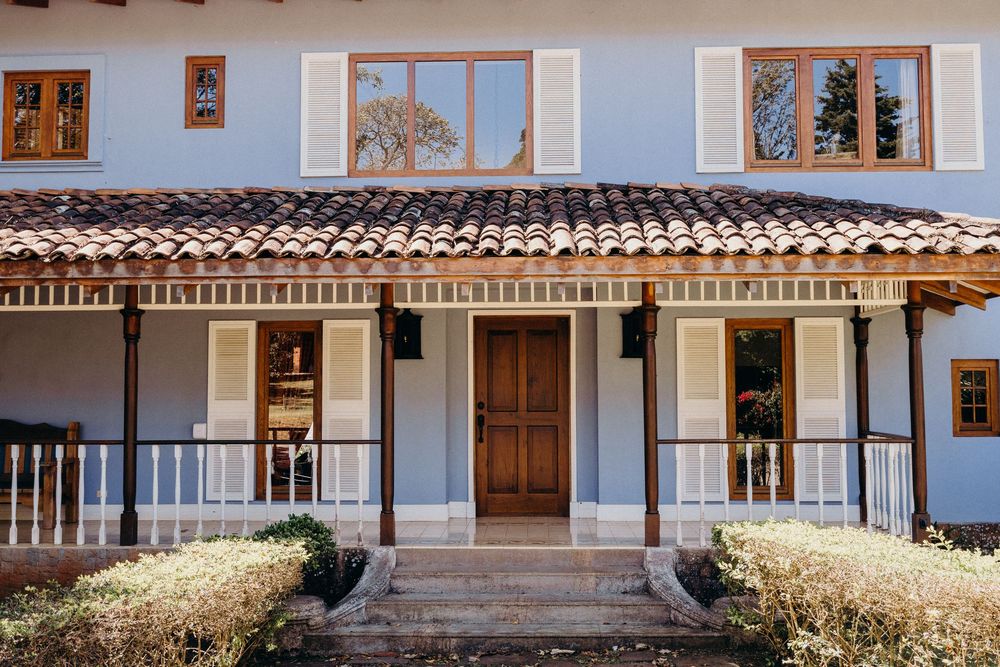 Elegant colonial-style home with terracotta roof, wooden door, shuttered windows, and manicured shrubs on a sunny day.