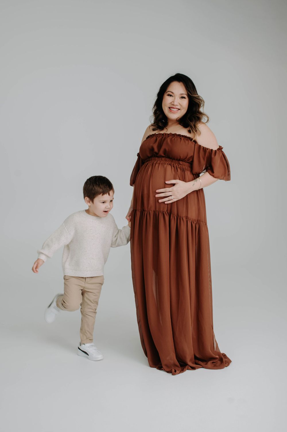 A maternity portrait showing a woman in brown dress with a child in studio setting.