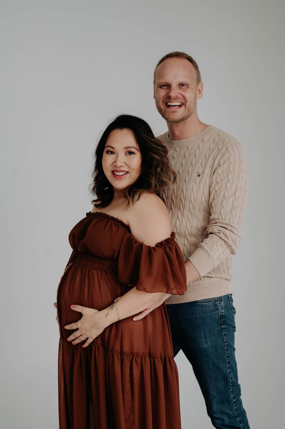Maternity portrait featuring couple in brown dress and beige sweater against studio backdrop.