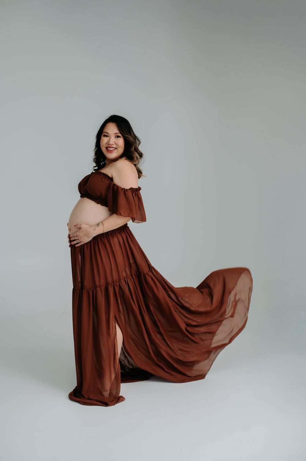 Expectant mother in flowing brown off-shoulder dress poses elegantly against gray backdrop.
