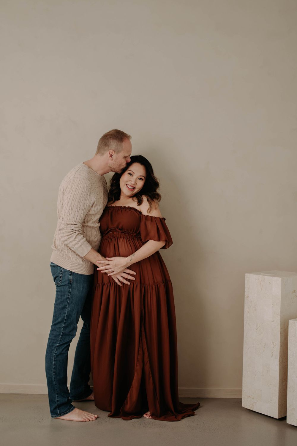 Intimate maternity portrait of couple in brown dress and beige sweater against neutral wall.