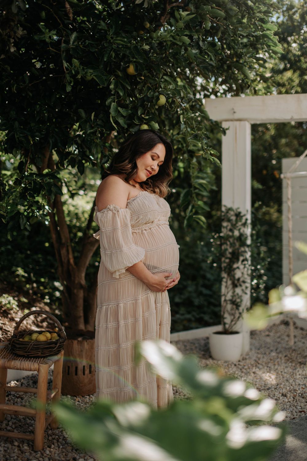 Outdoor maternity photo of pregnant woman in lace dress standing in garden setting.