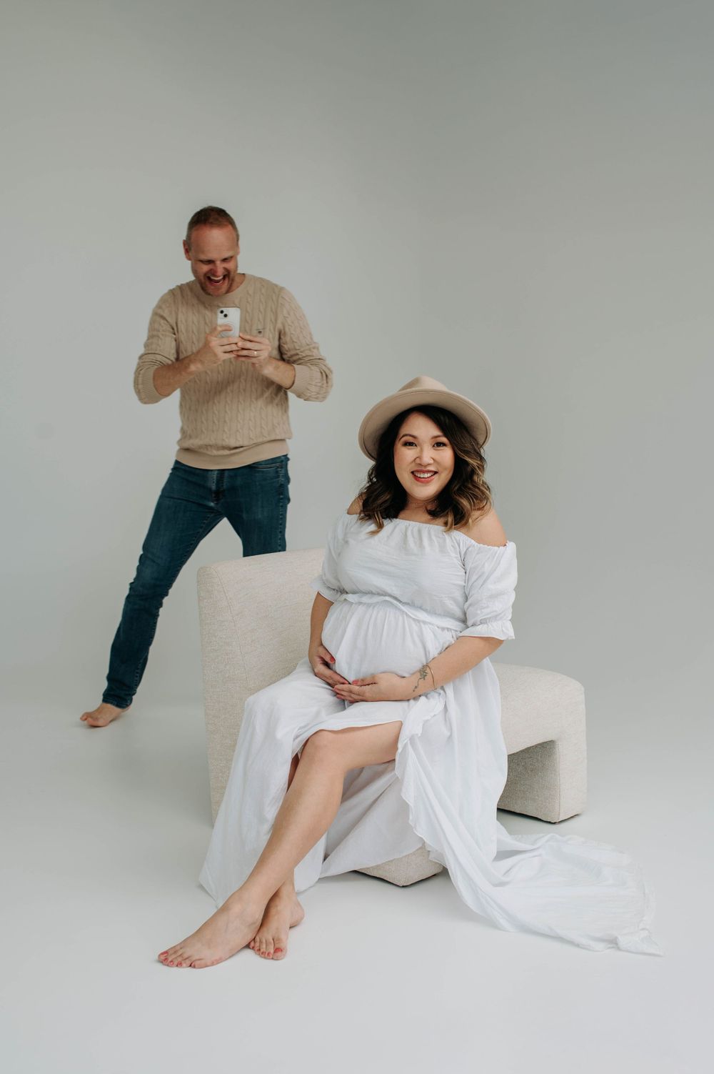 Playful maternity photo with person in beige sweater behind pregnant woman in white dress.
