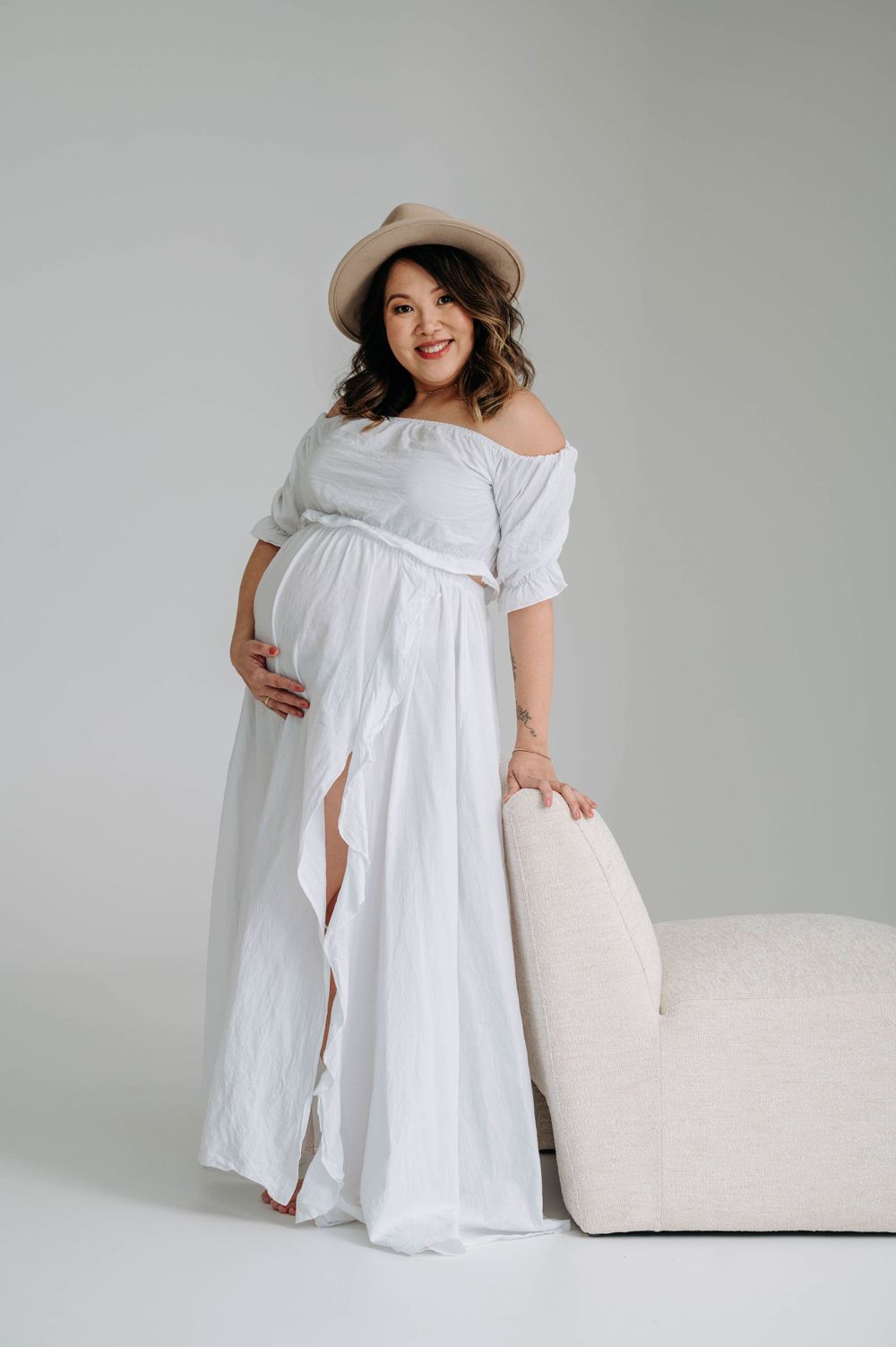 Pregnant woman in white off-shoulder dress and hat poses beside cream colored couch.