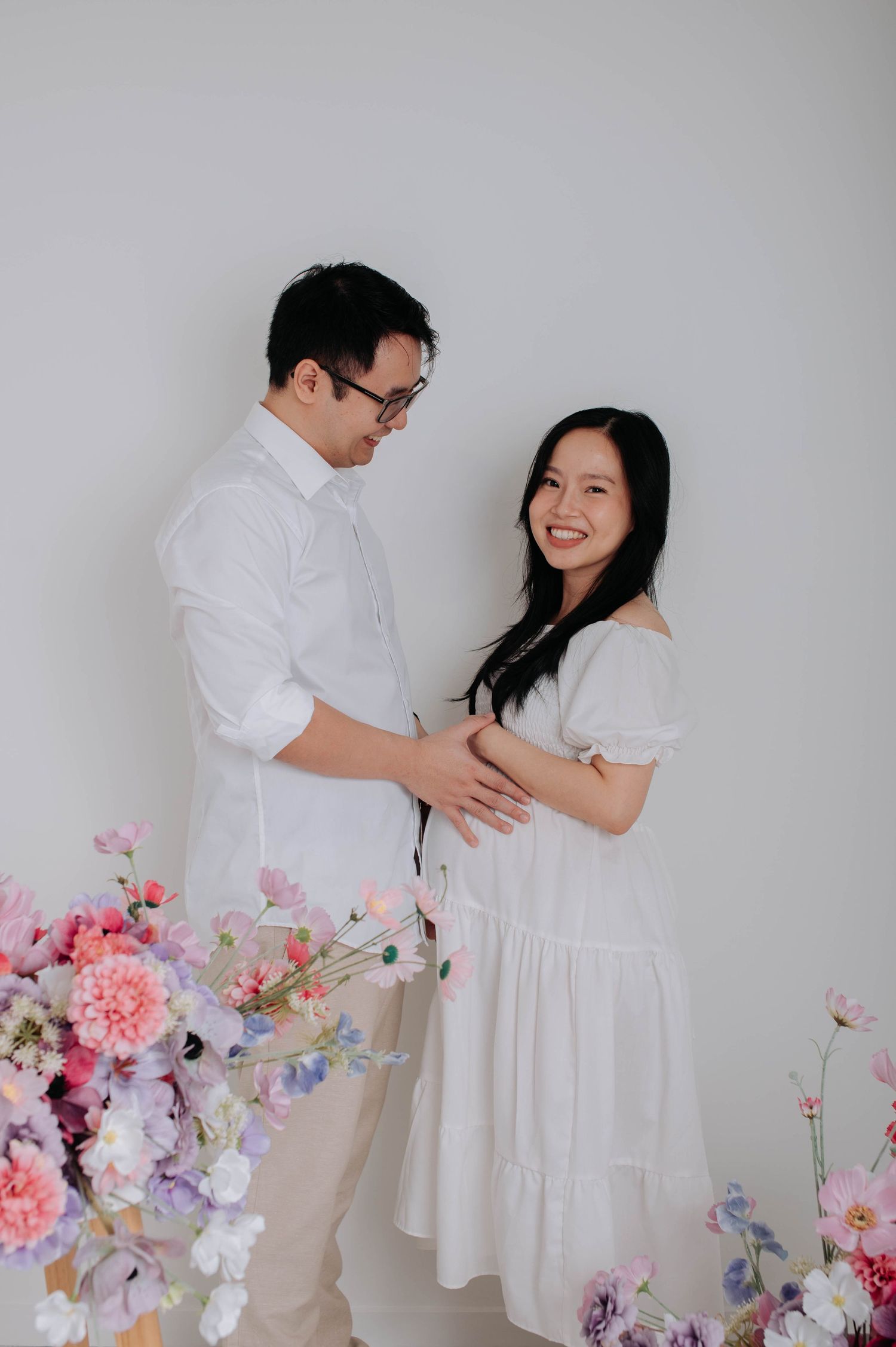 Couple in white outfits pose with floral decorations during pregnancy photoshoot.