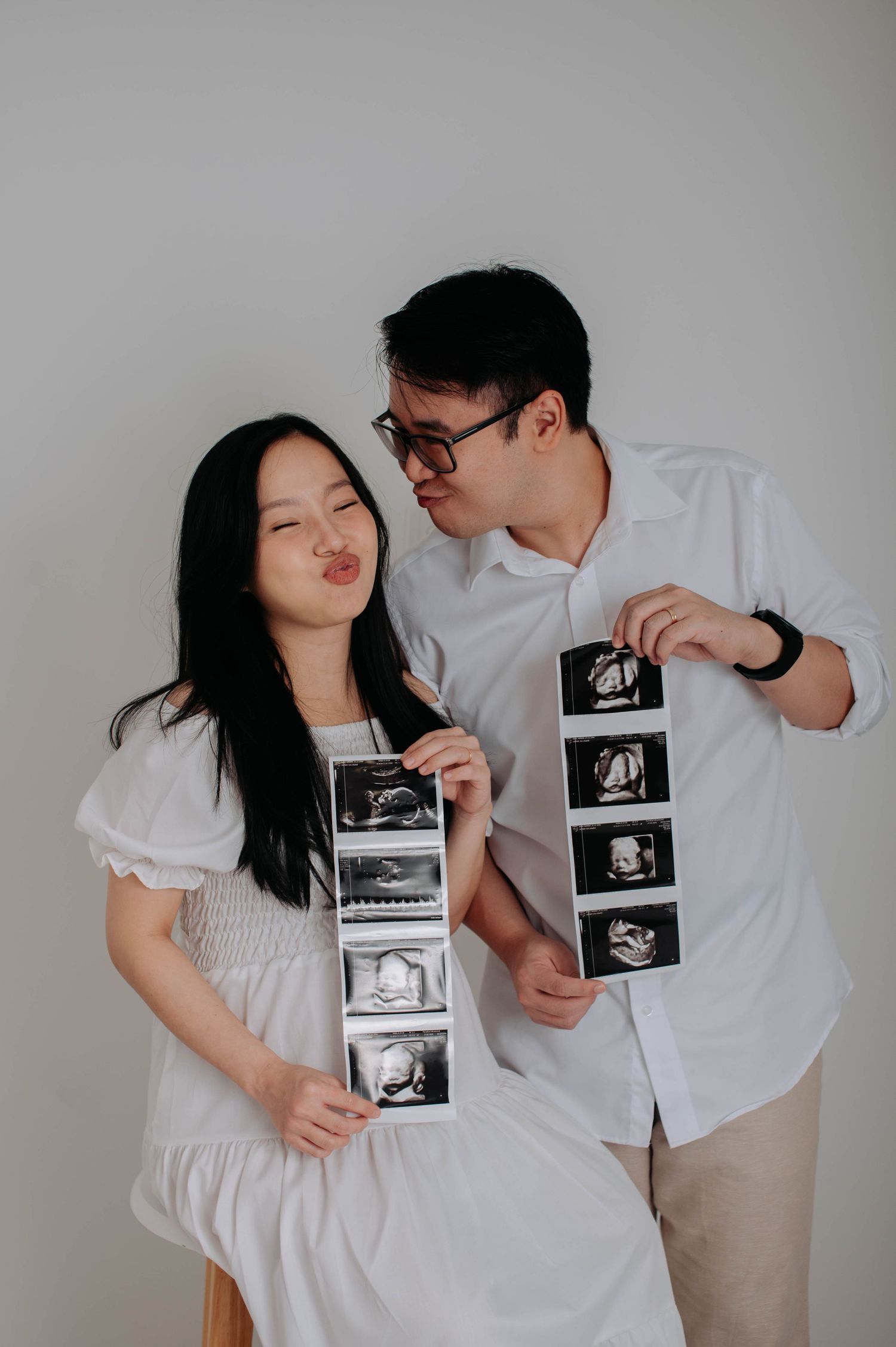 Expectant couple holding ultrasound photos while smiling against grey background.