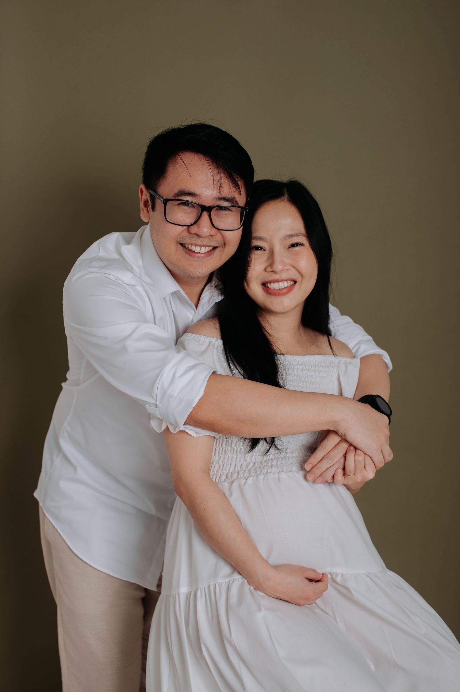 Maternity portrait of a couple in white attire embracing against a brown background.