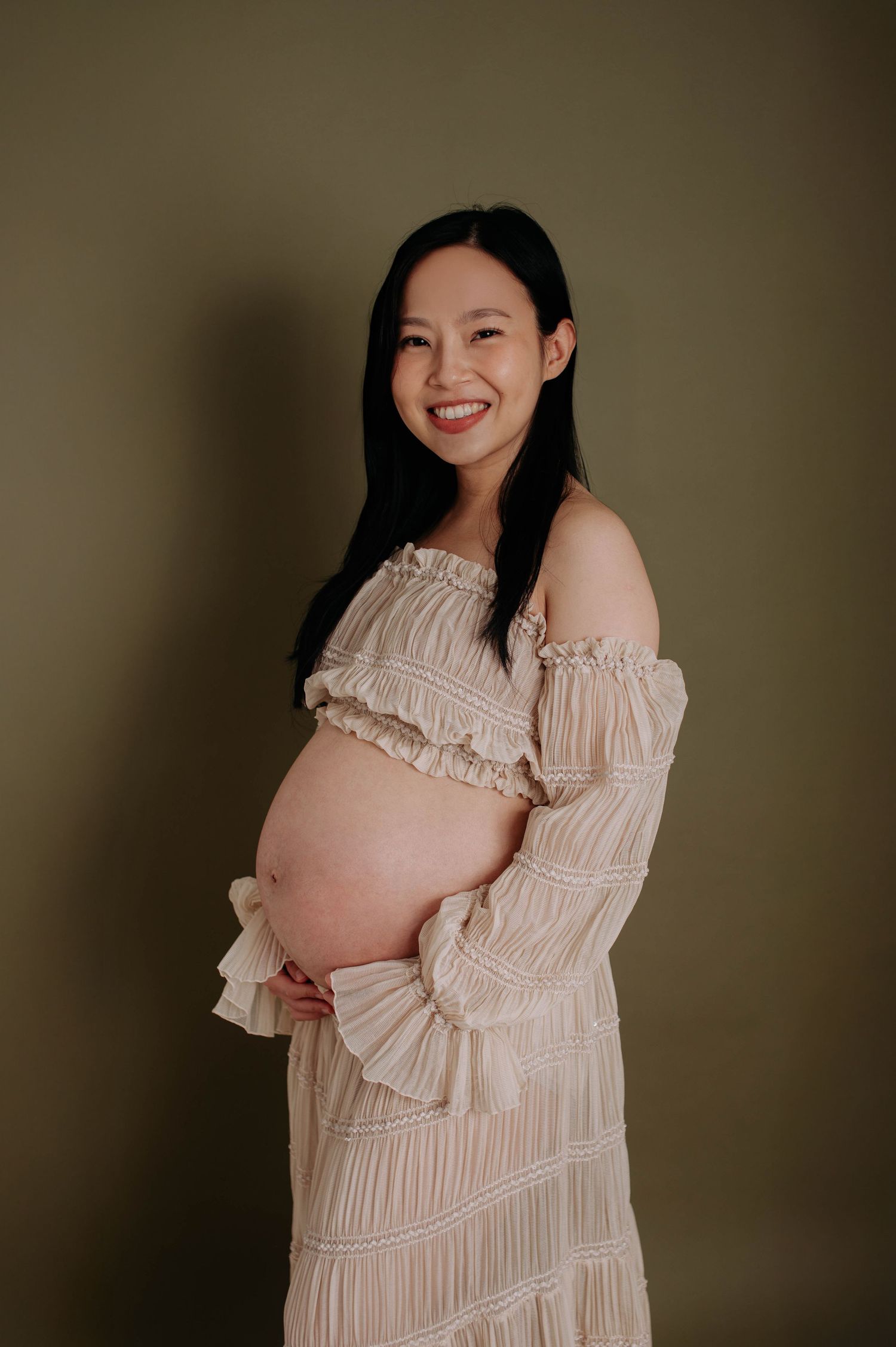 Pregnant woman in off-shoulder cream dress smiling during maternity photoshoot.
