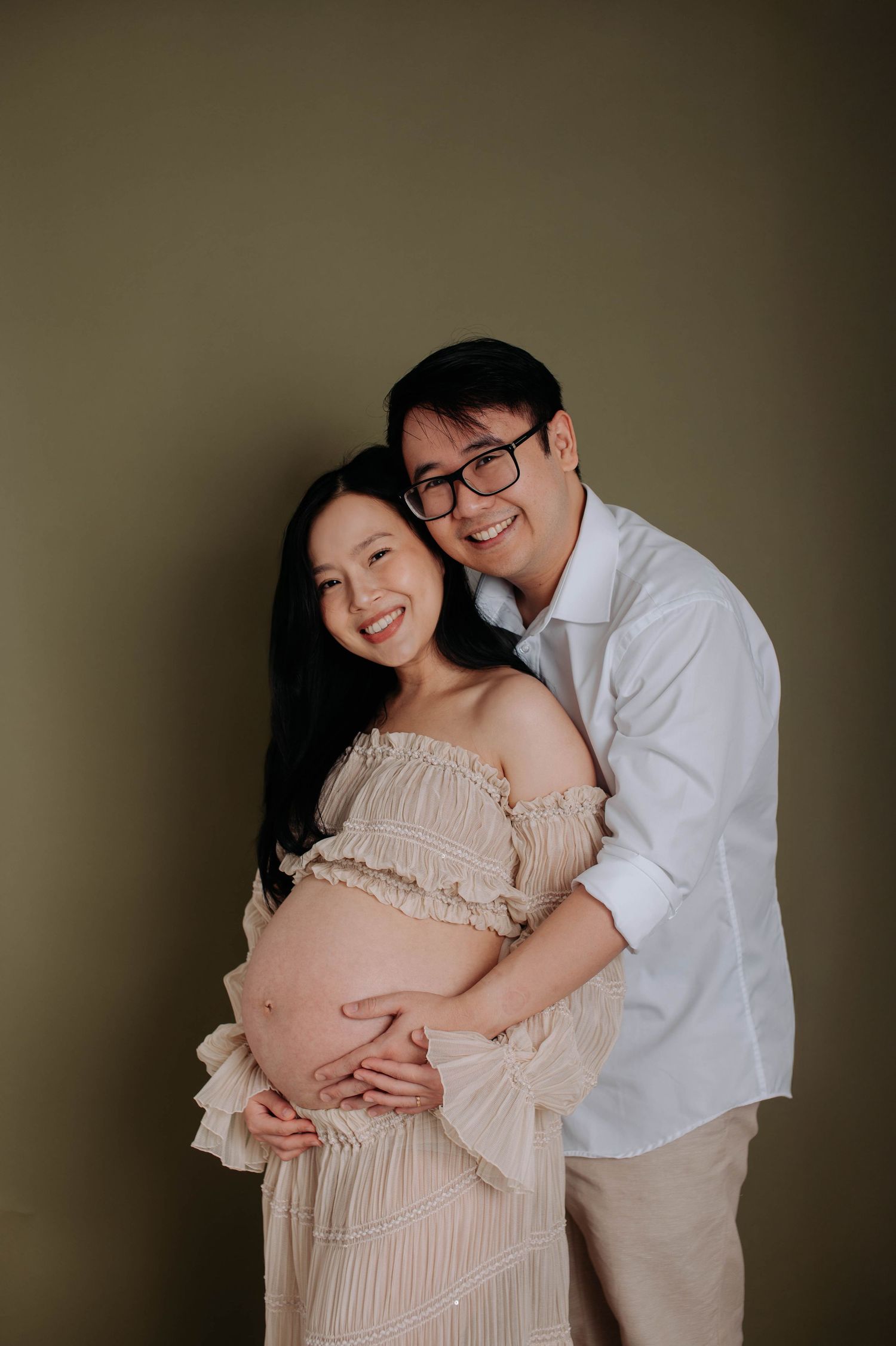 Maternity photo of couple in light colored outfits embracing baby bump against brown backdrop.