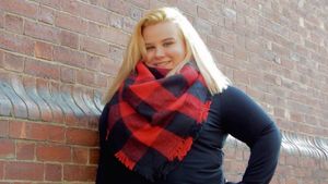 Person wearing a black sweater and red plaid scarf poses against brick wall.