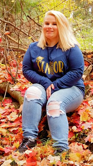 Person in navy sweatshirt sitting on log surrounded by autumn leaves.