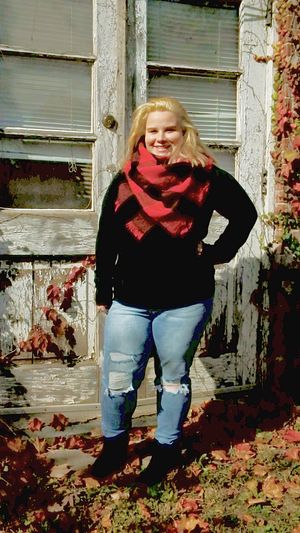 Person in black sweater and red scarf poses by weathered door with fall foliage.