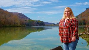 Person in plaid shirt stands by scenic lake with mountains in background.