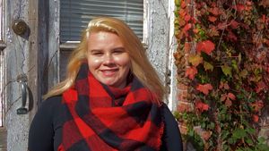 Person wearing red and black plaid scarf smiles by white door frame.