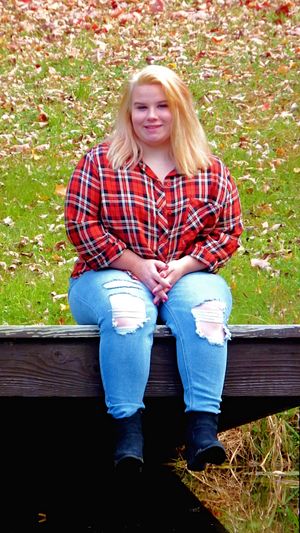 Person in red plaid shirt and ripped jeans sitting on wooden bench surrounded by fall leaves.