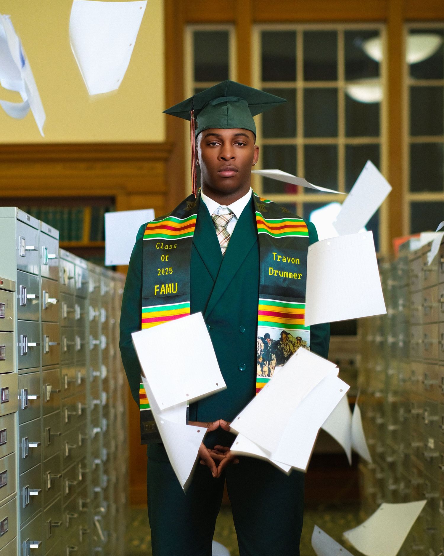 Graduate in green cap and gown with decorative stole throwing papers in hallway.