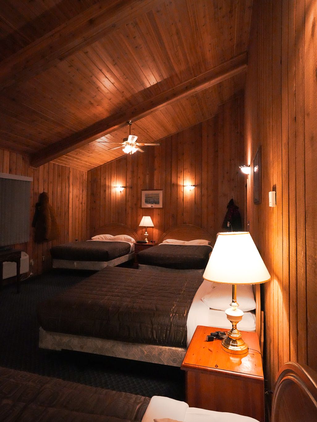 Rustic lodge bedroom with wood-paneled walls, multiple beds, and warm lighting fixtures.