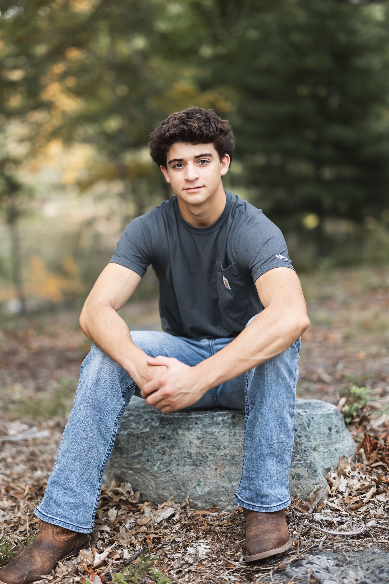 Handsome teenage boy in gray t-shirt and jeans sitting on a rock in an autumn forest setting.