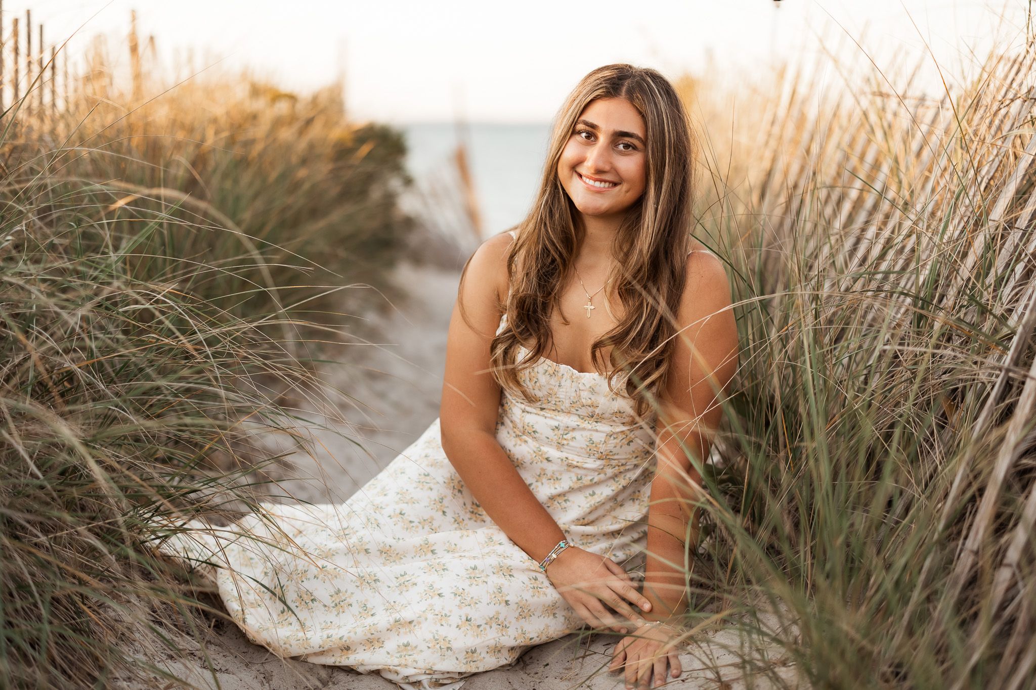 A smiling young woman in a floral dress sits among beach grass dunes during a golden hour senior portrait.