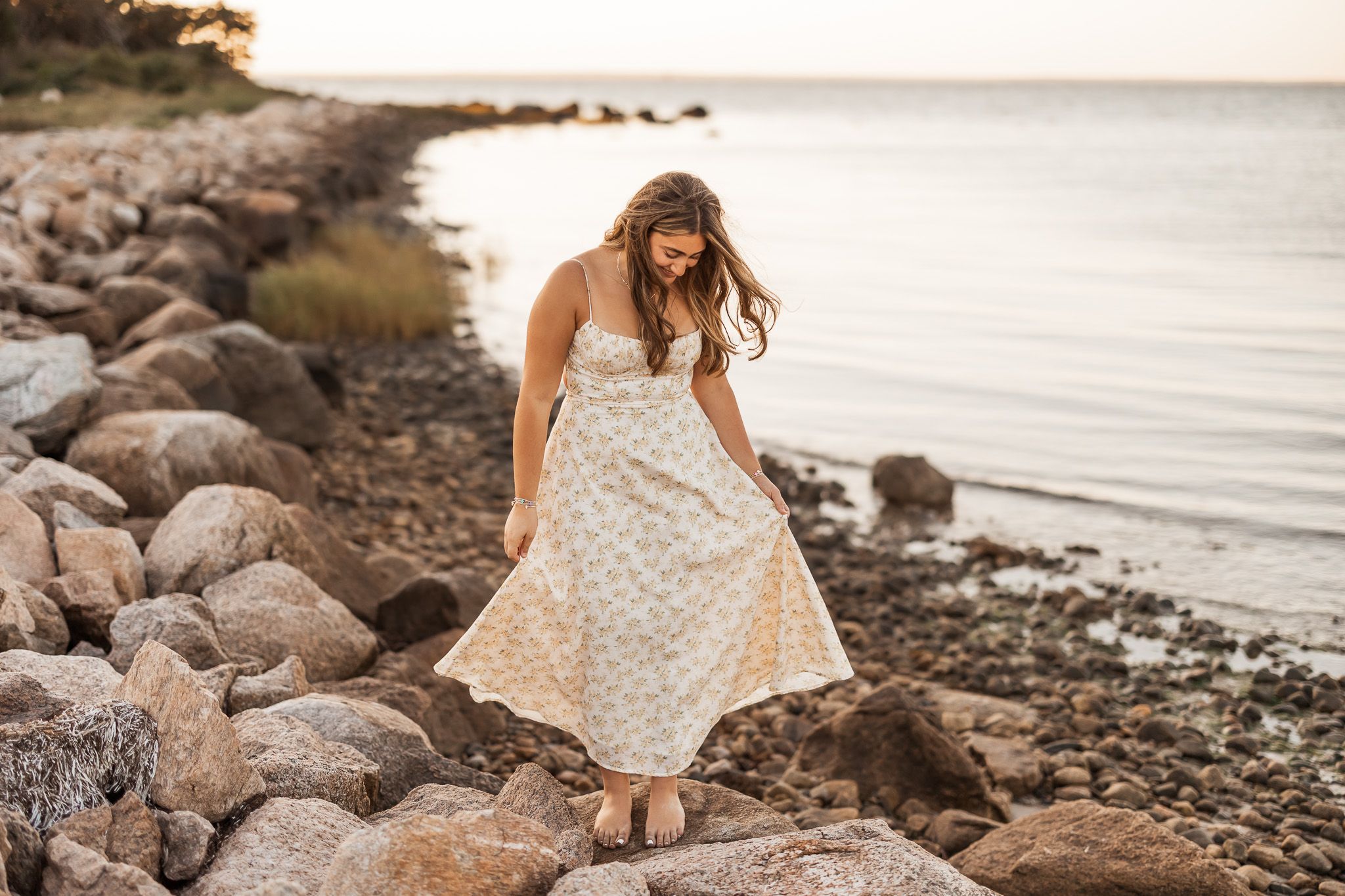 A young woman in a floral maxi dress walks along a rocky shoreline at golden hour sunset.