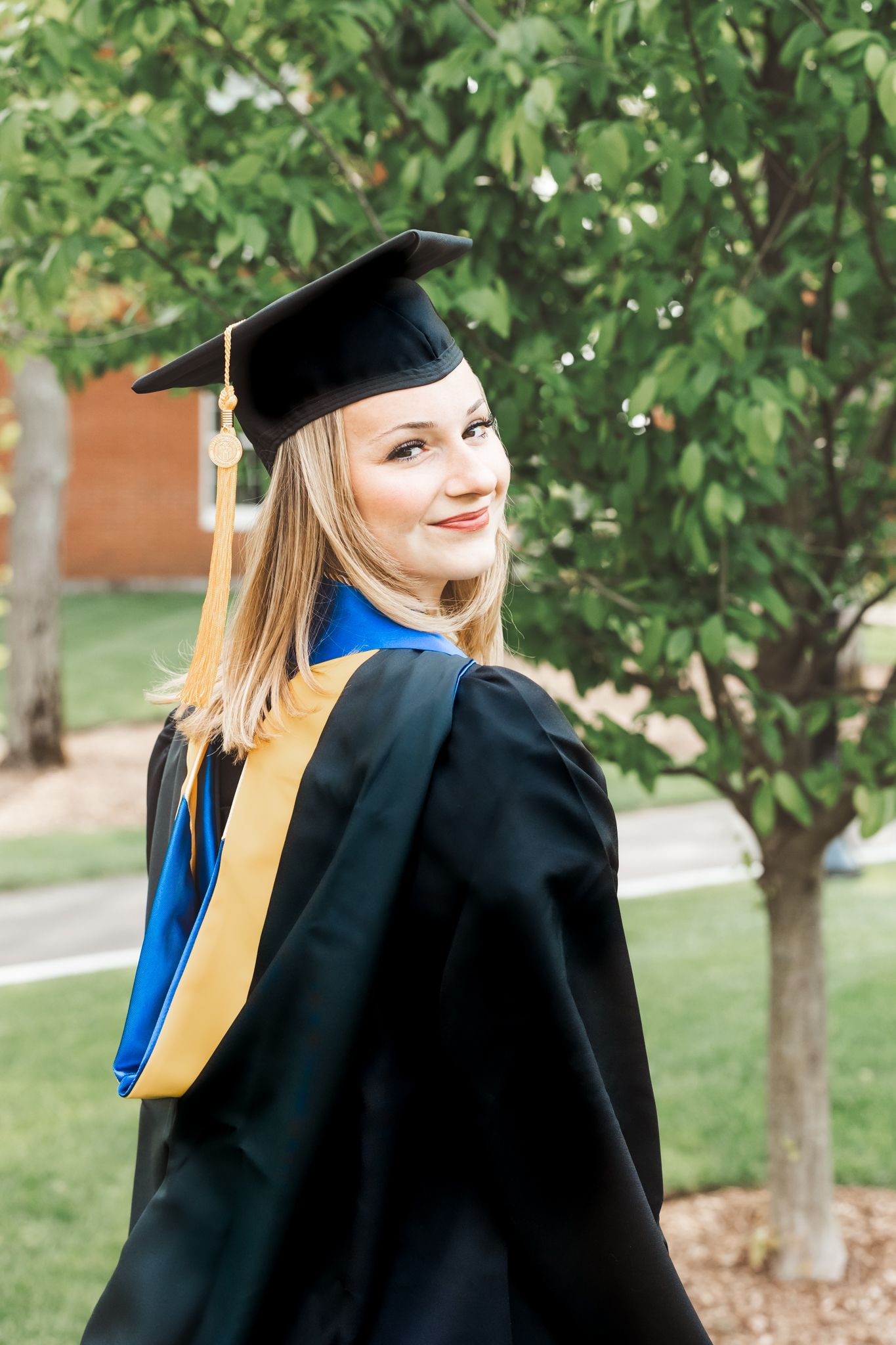 Smiling female graduate in black cap and gown with blue and gold regalia outdoors near trees.