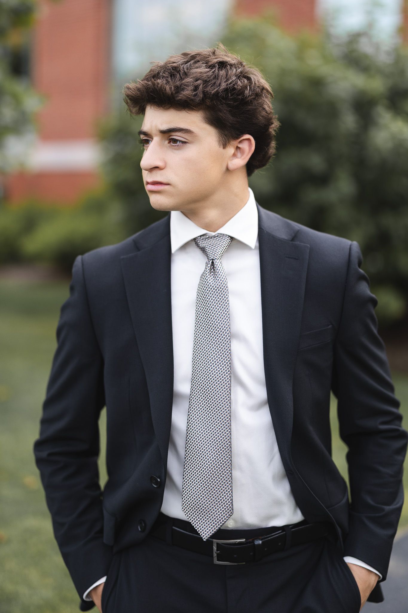 Stylish young man in dark suit and tie standing outdoors with hands in pockets, looking away.