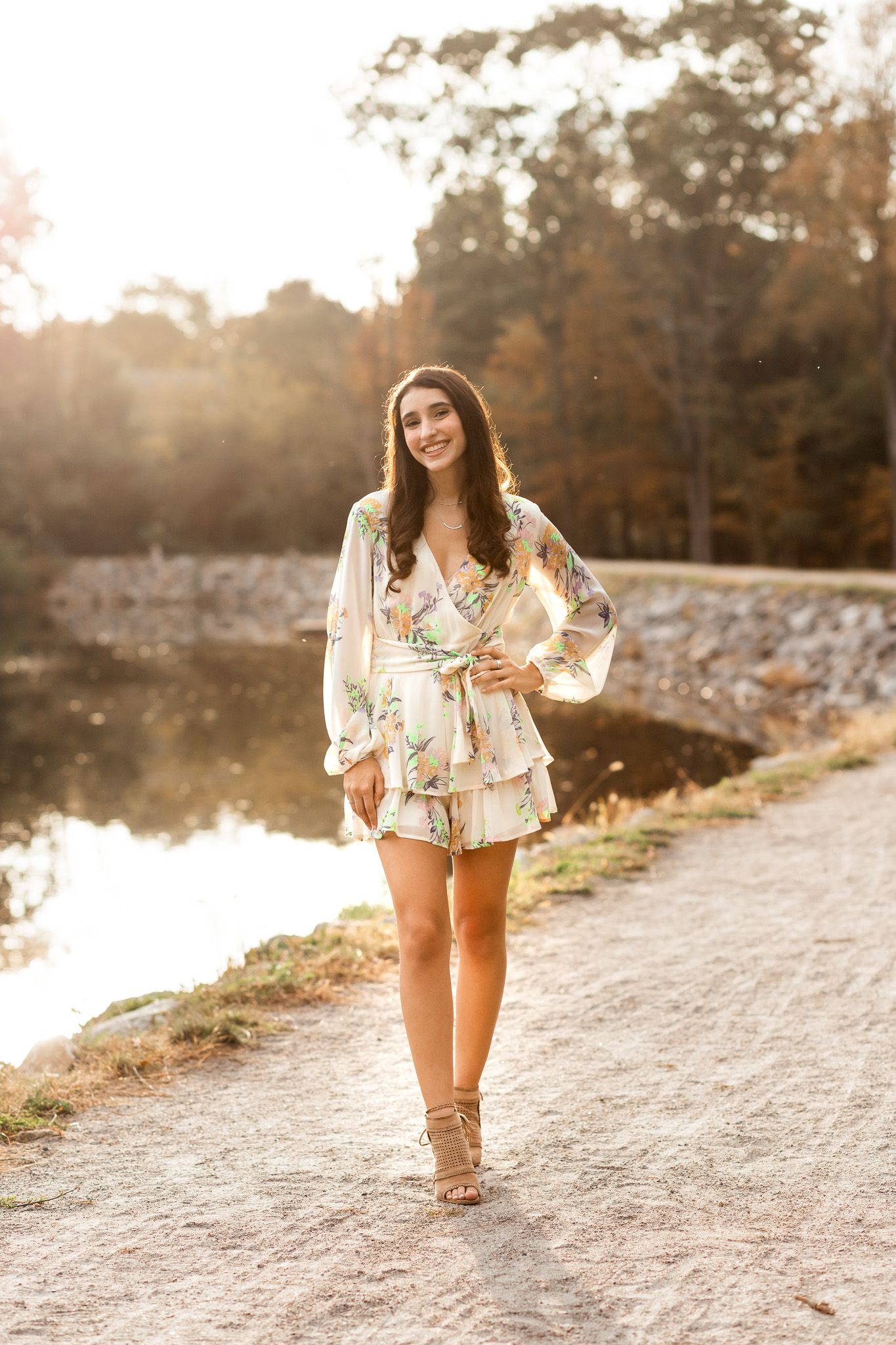 Smiling brunette woman in floral romper walking along scenic lakeside path during golden hour.