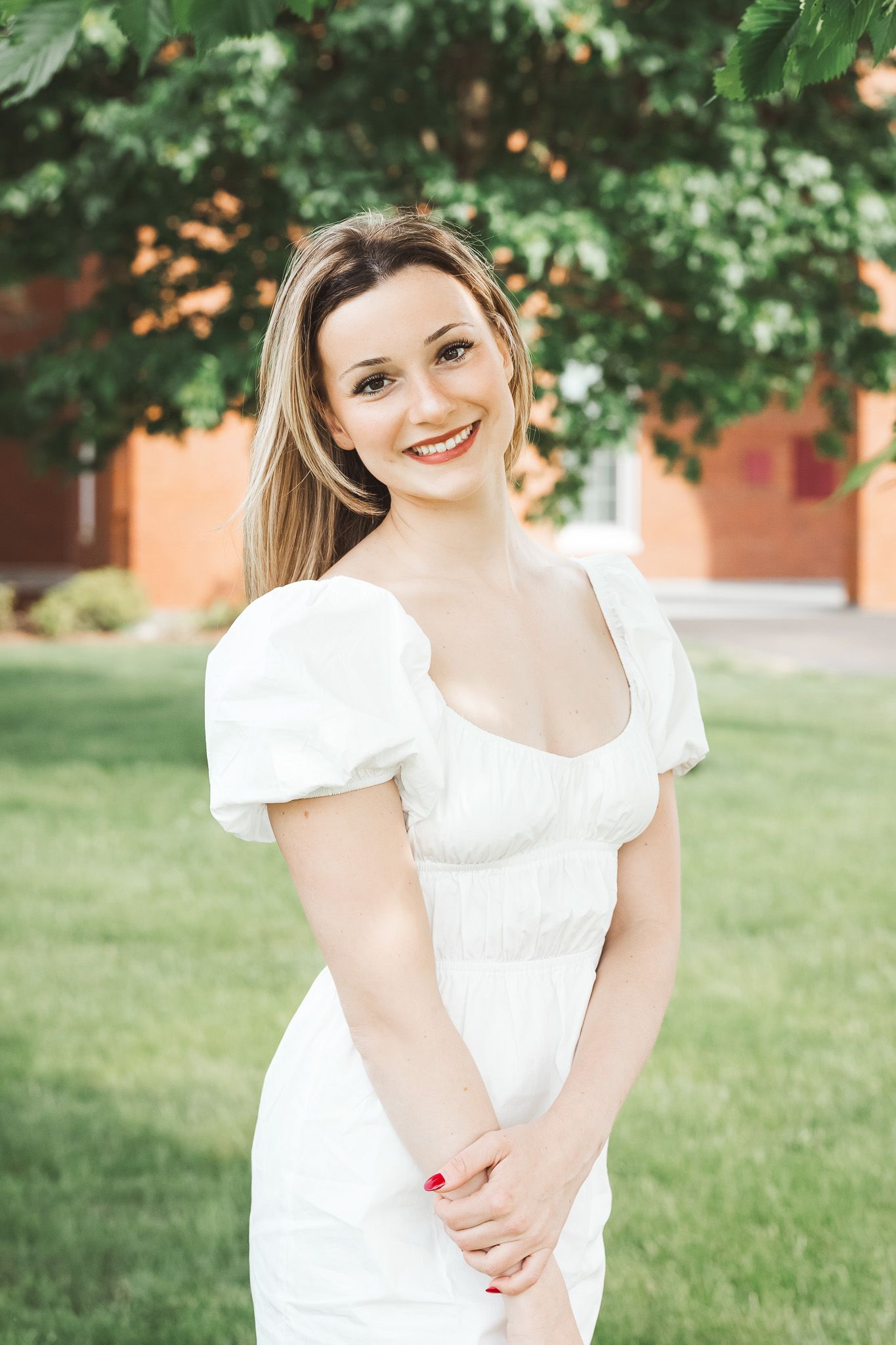 Smiling young woman in white puff-sleeve dress standing outdoors on green lawn near brick building.