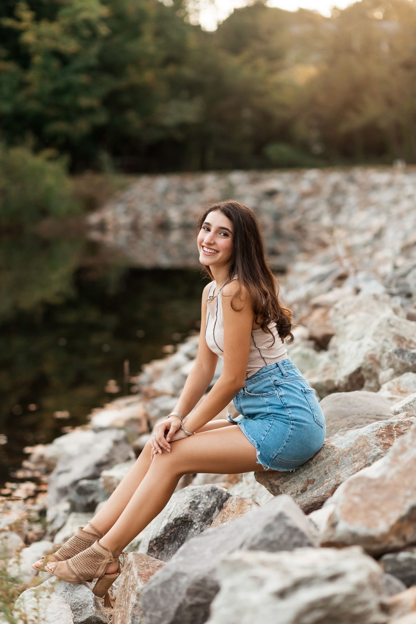 Young woman in denim shorts sitting on rocks by a river, smiling at golden hour.