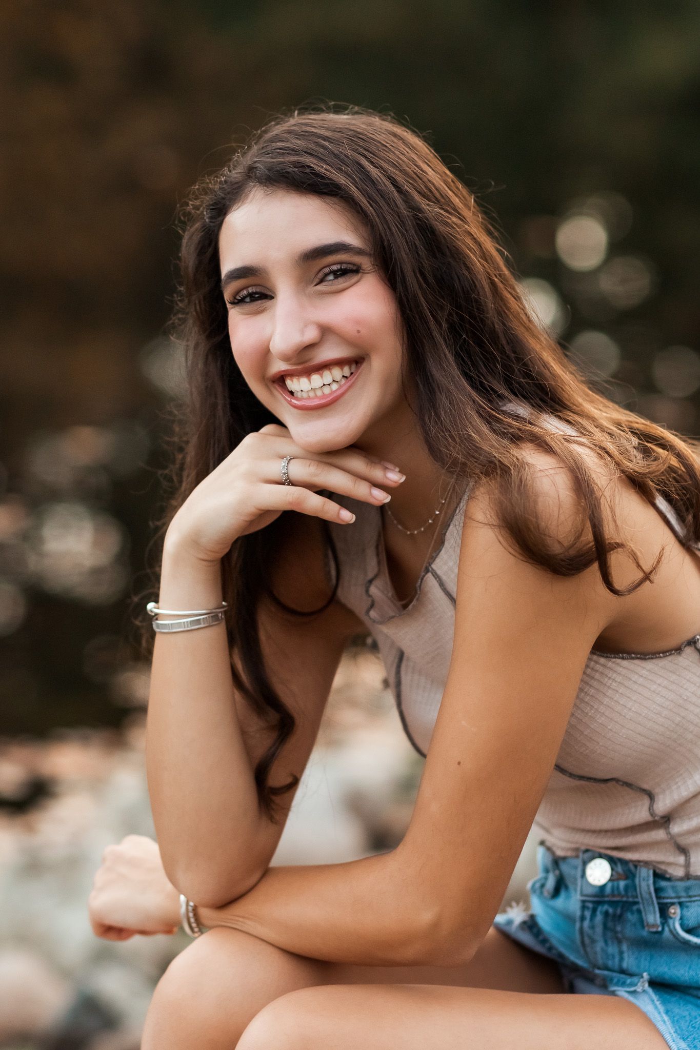 Smiling teenage girl with long brown hair posing outdoors wearing a tank top and denim skirt.