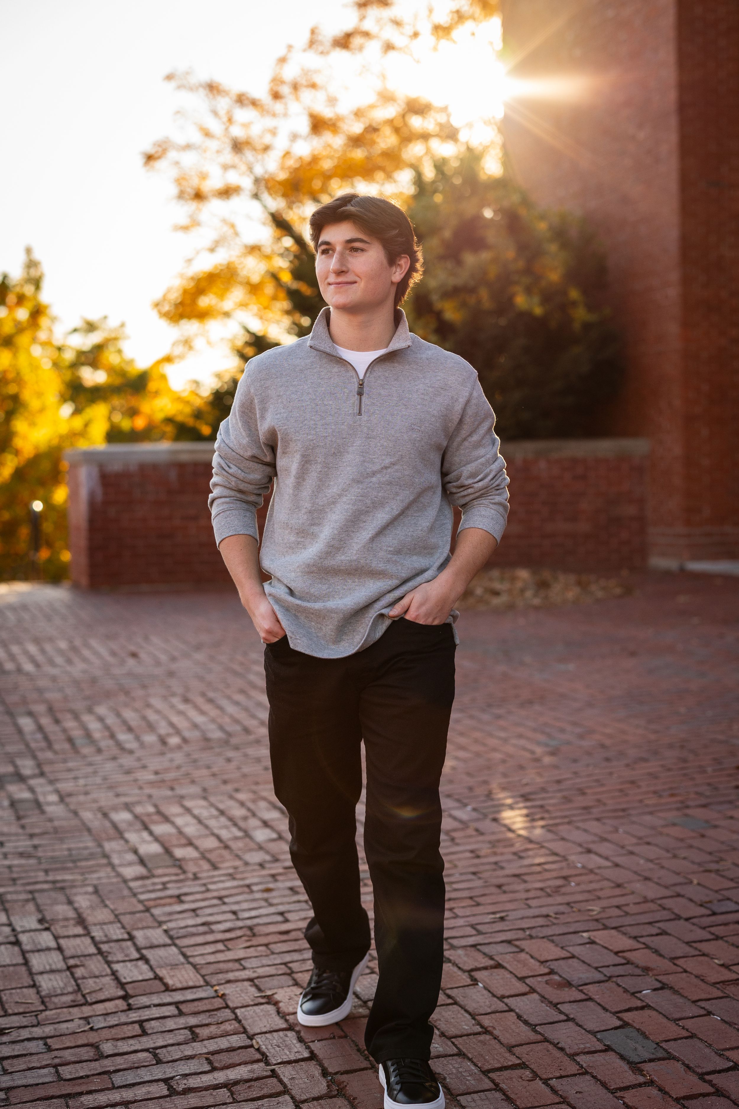 Young man in gray quarter-zip sweater walking on brick path at sunset.
