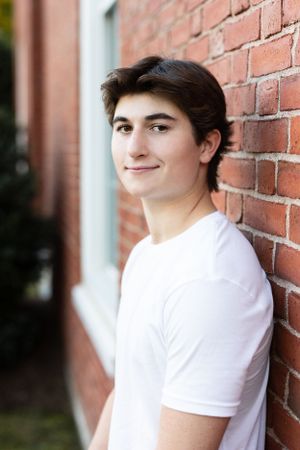 Teen boy in white t-shirt leaning against red brick wall, smiling outdoors.