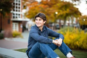 A senior portrait of a young man in a blue shirt sitting outdoors on steps surrounded by fall foliage.