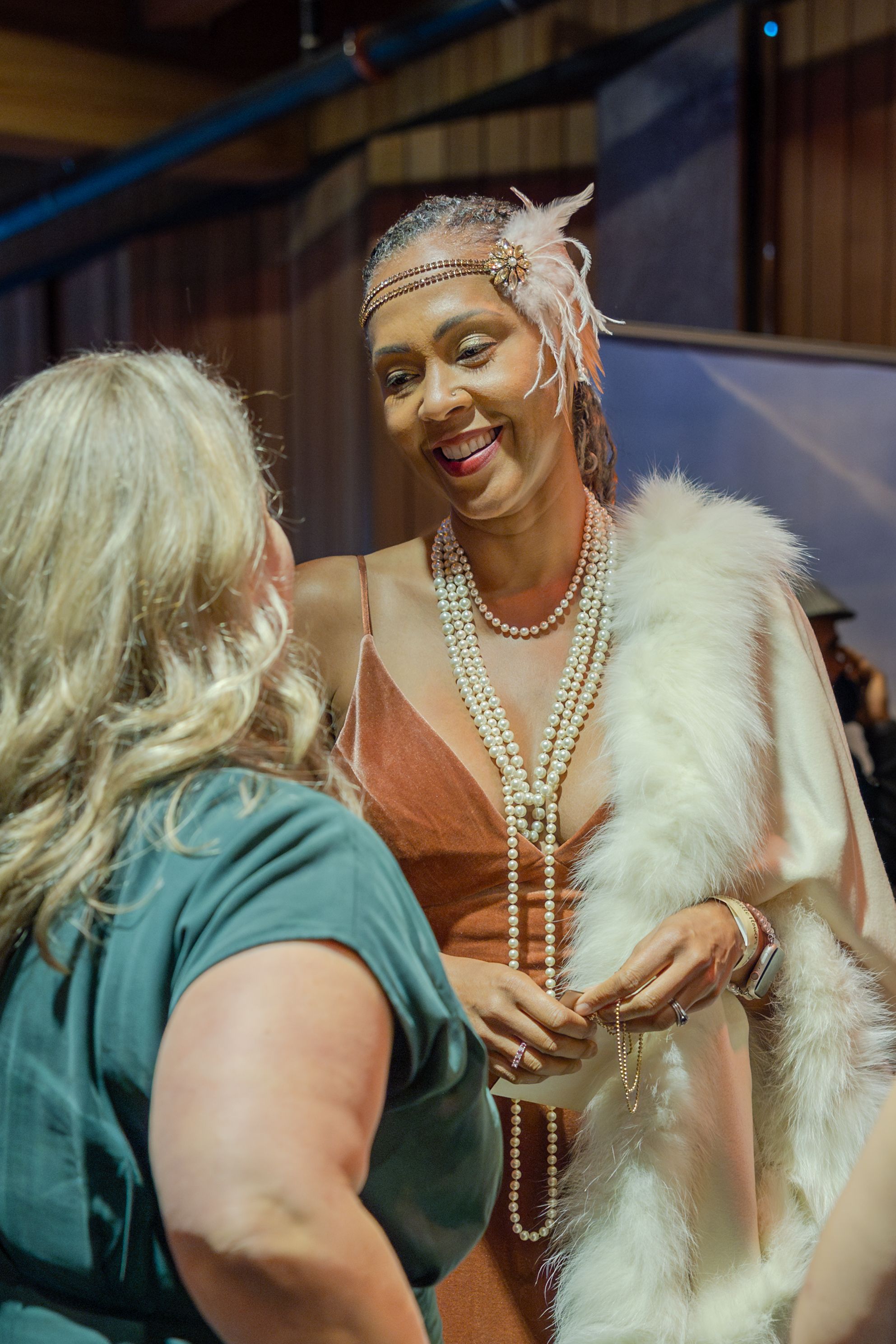 Woman in 1920s flapper costume with pearl necklaces and feather headband smiling at another woman at a party.