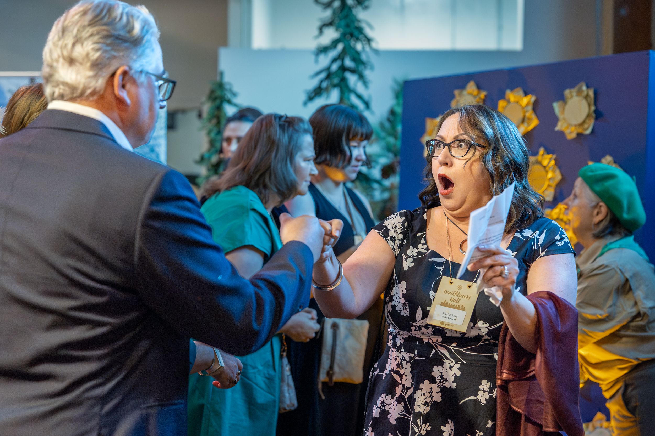 Woman reacts with surprise while greeting a man at a professional networking event with attendees in background.