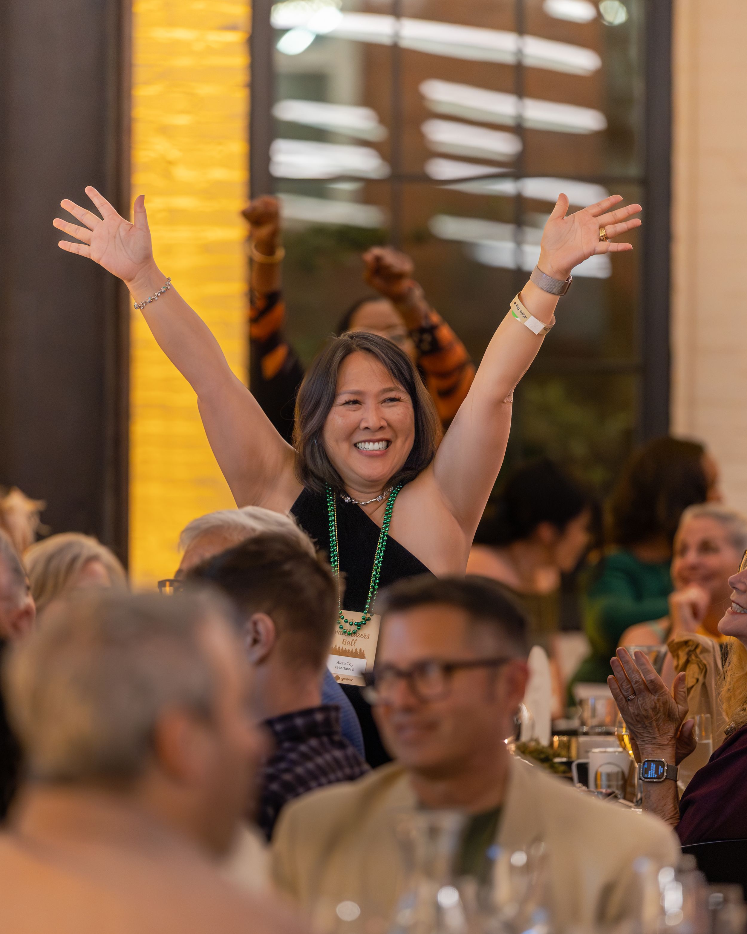 Woman raises arms in celebration at a conference dinner event, smiling joyfully among seated guests.