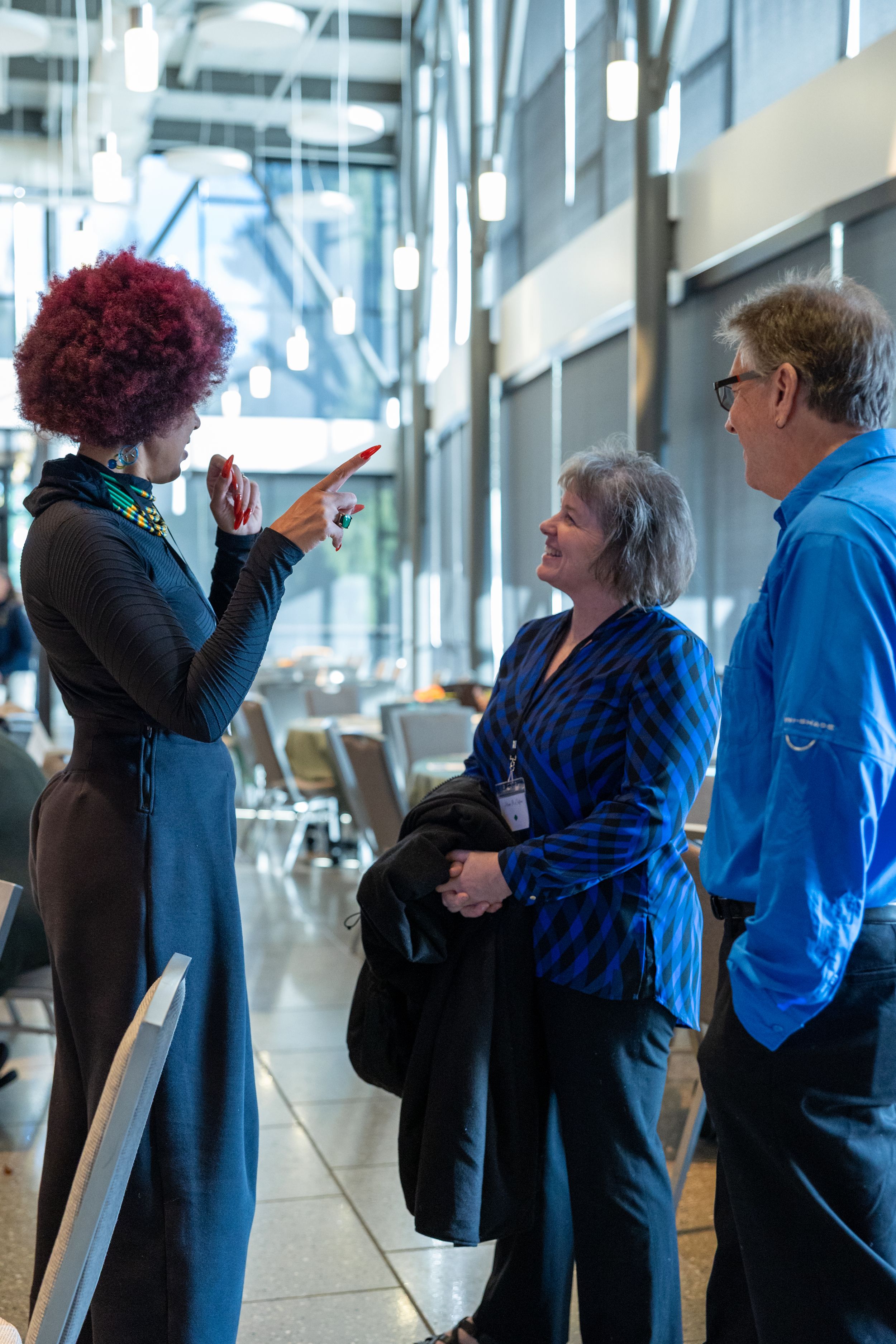 Three professionals conversing in a modern building, woman with red curly hair pointing while explaining to colleagues.