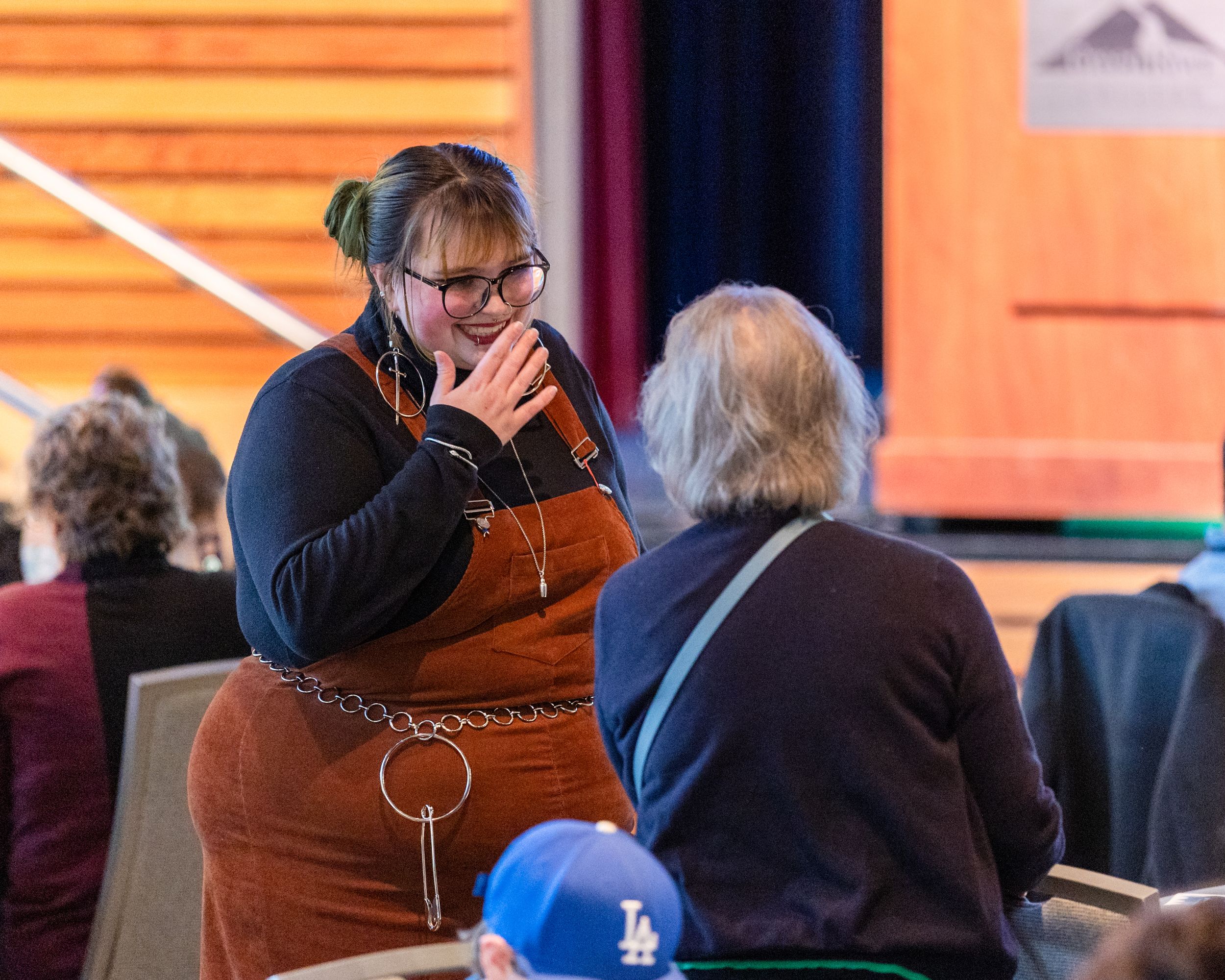 Woman in orange overalls and headphones gestures while conversing with seated elderly person at indoor event.