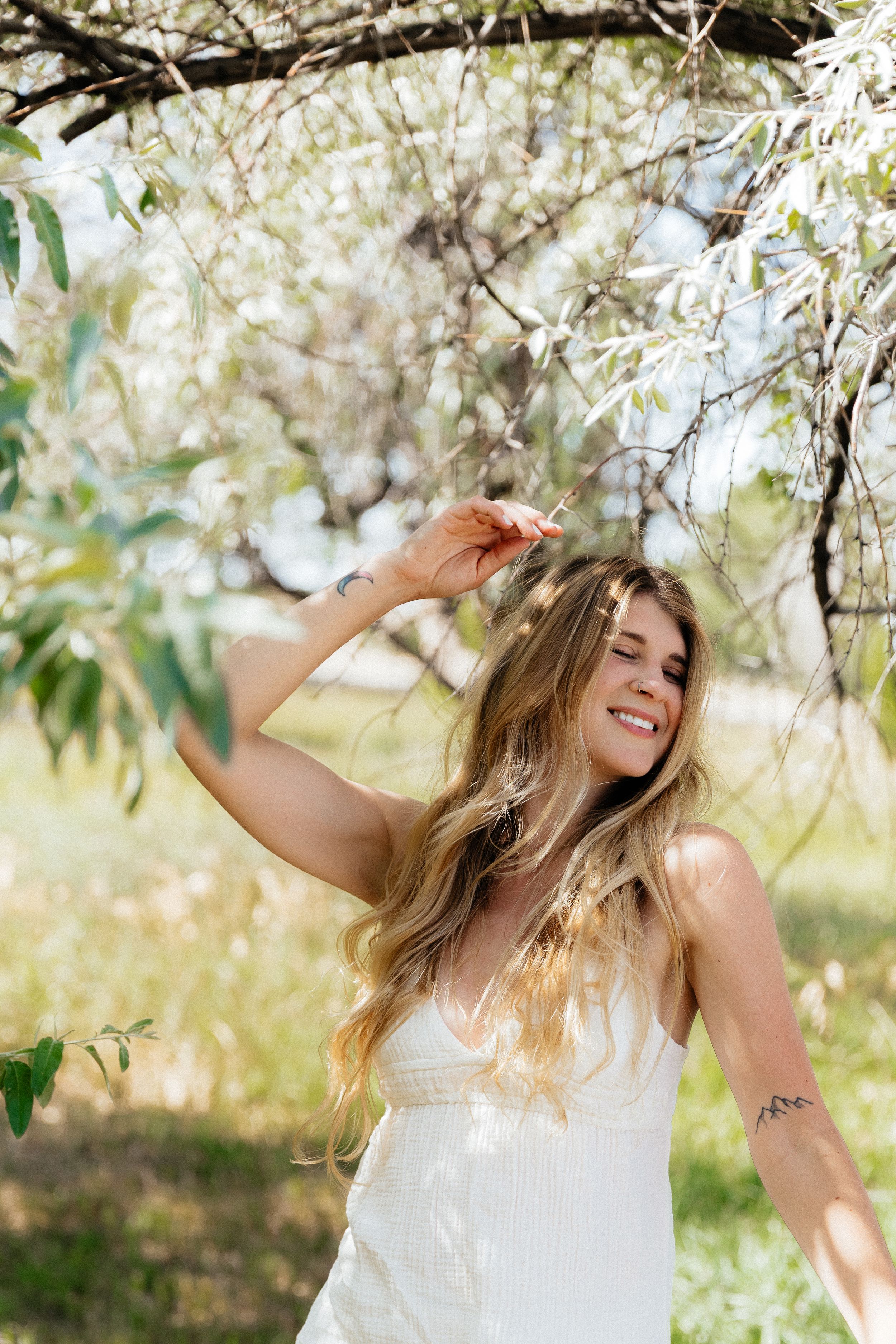 A smiling woman with long blonde hair in a white sundress poses among blooming spring tree branches outdoors.