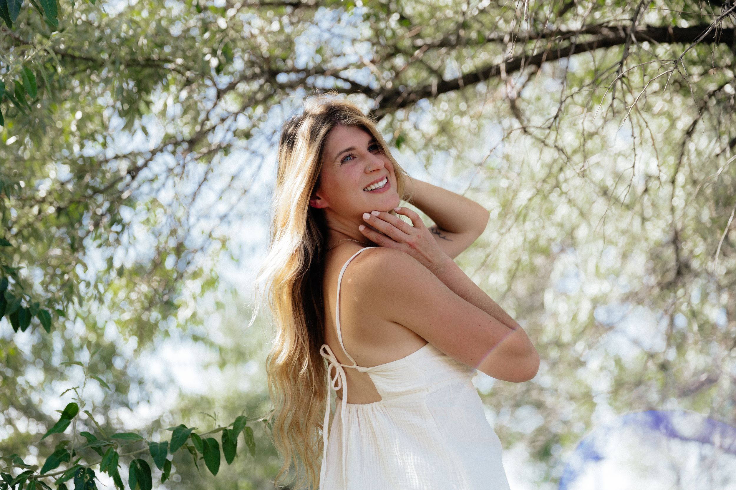 Young woman with long blonde hair in white dress smiling outdoors under green tree branches.