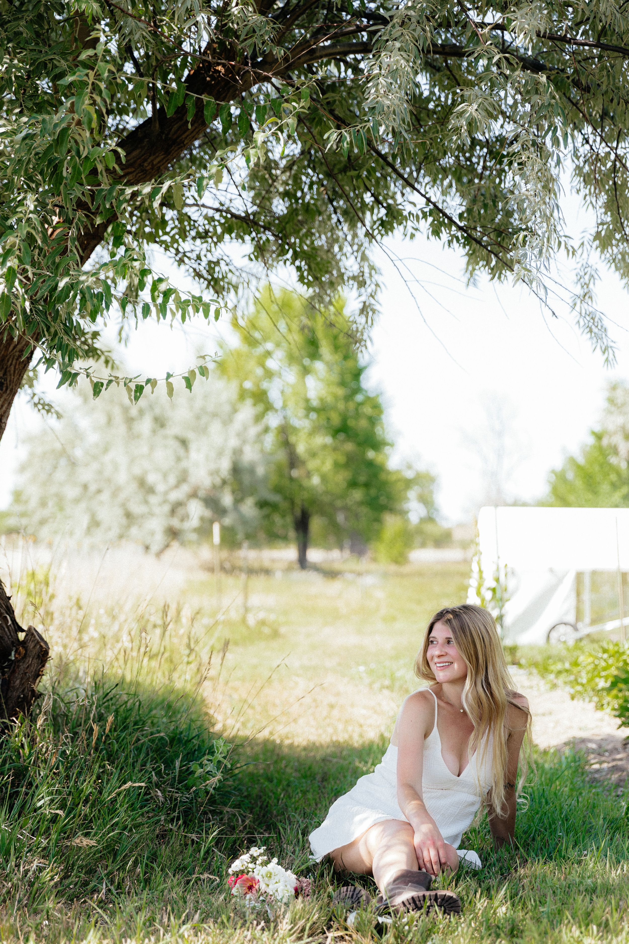Woman in white dress sitting under a tree on green grass, smiling and looking up in a serene outdoor setting.