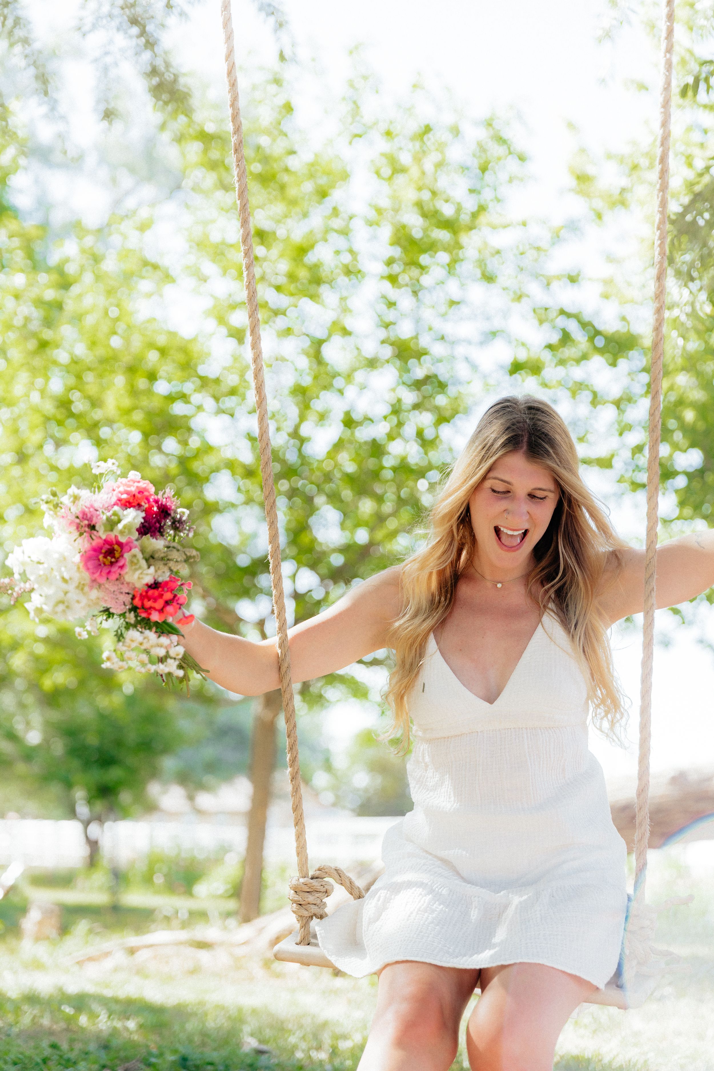 Joyful blonde woman in white dress holding pink flowers while swinging on a rope swing outdoors.