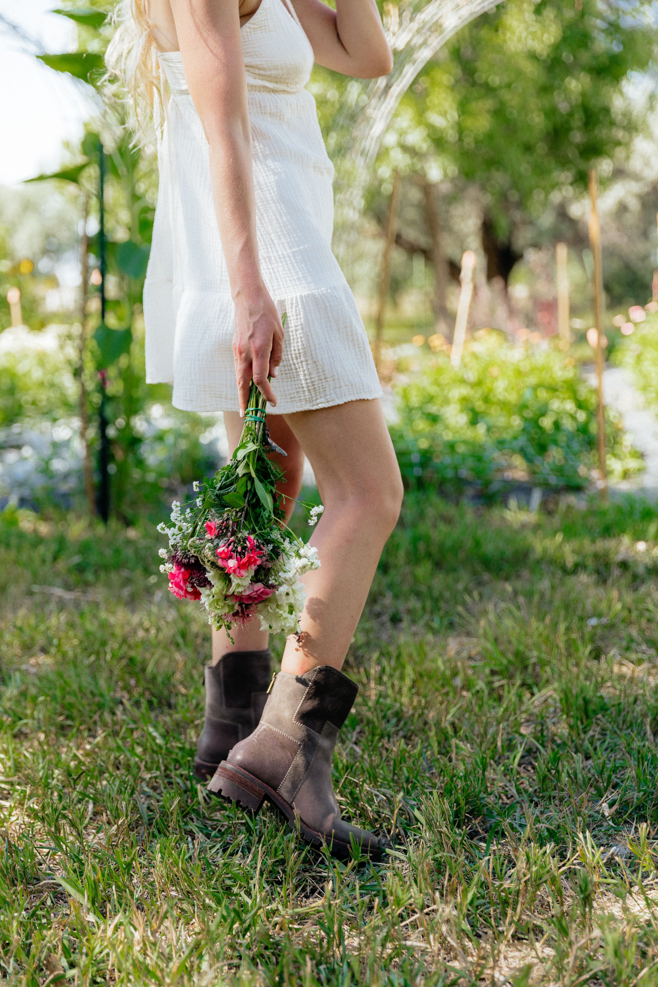 Woman in white dress and brown boots holding a bouquet of wildflowers while standing on green grass outdoors.
