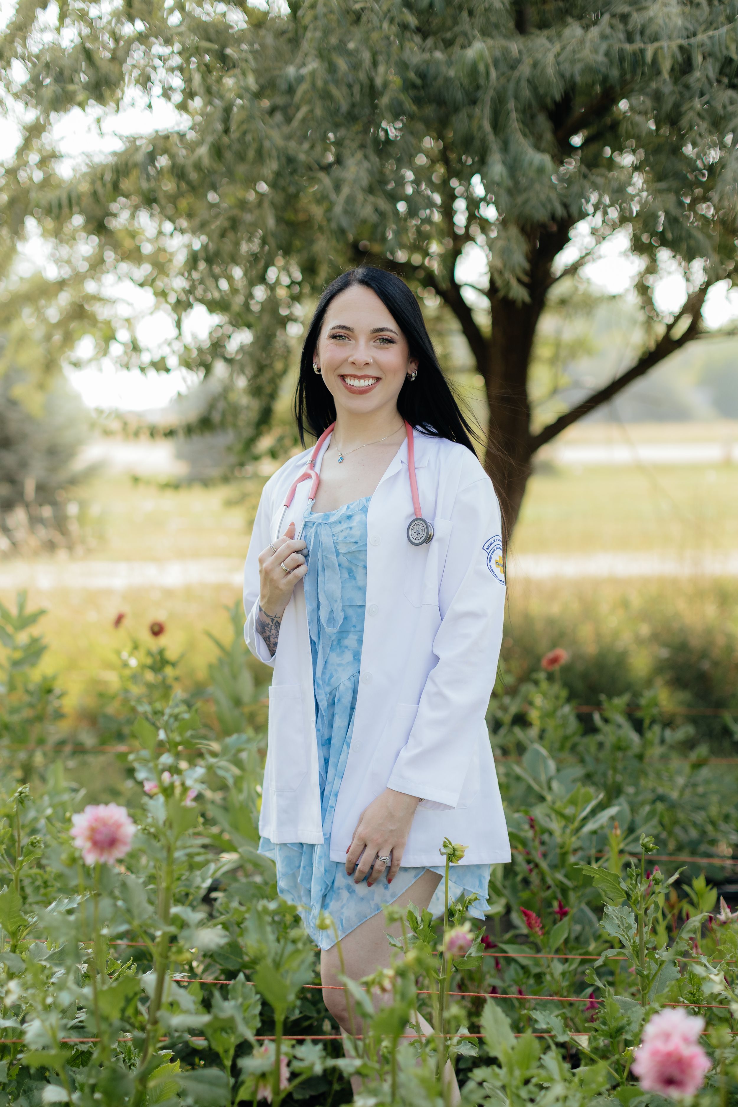 Smiling female doctor in white lab coat with pink stethoscope standing outdoors among flowers and greenery.